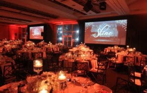 In the shadowy corners of a grand banquet hall, a league of round tables emerges, each adorned with vibrant floral centerpieces and flickering candles, ready to host an unforgettable gathering. Towering above, colossal screens blaze with the emblematic Shine insignia, casting an inspiring glow across the room. A masterful Michigan wedding DJ orchestrates an electrifying atmosphere that charges through the air like lightning. Meanwhile, a photo booth rental stands as a fortress of fun, capturing heroic moments for every guest brave enough to step inside and immortalize their night in snapshots of glory. :The Event Heroes at Black Tie Productions