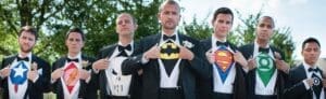 Black Tie Productions: Seven men in tuxedos stand outdoors, revealing superhero logos under their shirts—Captain America, Flash, Punisher, Batman, Superman, Green Lantern. Perfect for a fun Michigan wedding entertainment scene with a vibrant twist. Trees and a clear sky set the perfect backdrop. :Your Event Heroes