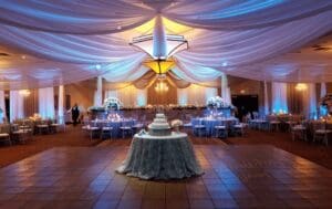 In the grand hall of elegance, where destiny meets design, a majestic wedding reception unfolds. The ceiling, draped like the cape of a guardian hero, watches over a central table showcasing a towering masterpiece—cake fit for champions. Around it, round tables stand ready to host noble guests in their joyous assembly. Blue uplighting bathes the scene in an ethereal glow, while floral arrangements add bursts of nature’s own superpower. Anchoring this realm is an expansive dance floor—space where dreams take flight and moments become legendary. For those seeking a photographic legacy worthy of the superheroes they are destined to be, Black Tie Productions swoops in with premium photo booth rental experiences beyond imagination!. :The Event Heroes at Black Tie Productions