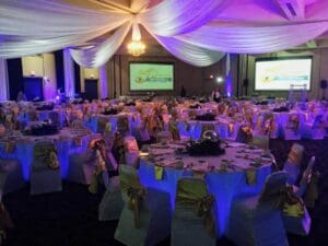 In a realm where celebrations reach legendary proportions, an extraordinary event unfolds within the grand banquet hall. The round tables stand as noble guardians, cloaked in pristine white and radiant gold. Celestial drapery cascades from above, transforming the ceiling into a tapestry of elegance. With an electric pulse, specialized lighting illuminates every corner with sophistication, casting shadows of heroic enchantment. Twin beacons—mighty screens—proudly showcase a glorious emblem: the mark of a 50th-anniversary triumph. To craft your own unforgettable saga within this shimmering fortress of festivity, summon Black Tie Productions! Unleash their power to conjure instant price quotes that promise unparalleled marvels at your event!. :The Event Heroes at Black Tie Productions