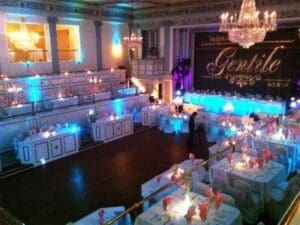 Black Tie Productions: A grand ballroom set for a wedding reception features round tables draped in white tablecloths with pink napkins. Chandeliers illuminate the room warmly, while soft blue and white uplighting enhances the elegant ambiance. A stage with a large banner awaits, promising top-tier party entertainment. :Your Event Heroes