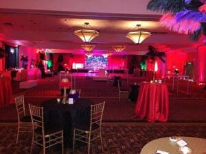 In the heart of the city, where heroes and legends gather, lies a ballroom transformed by the vibrant glow of red lights. Round tables stand proud, cloaked in dramatic black and powerful red, marking a realm of sophistication fit for champions. Overhead, chandeliers cast their radiant light like stars above Gotham. The stage is primed for an epic celebration orchestrated by none other than Black Tie Productions—masters in crafting unforgettable adventures. As twilight descends and the night unfurls its mysterious cloak, its time to immortalize memories: consider deploying a photo booth rental to capture every heroic moment under these hallowed chandeliers!. :The Event Heroes at Black Tie Productions