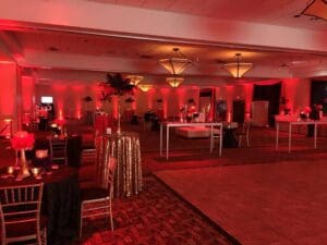 Amidst the buzzing cityscape stands an opulent event hall, draped in the heroic hues of crimson light. Majestic tables gleam beneath golden thrones, awaiting the gathering of eminent guests. Towering centerpieces rise like vigilant sentinels beneath a canopy of geometric beams that spread their radiant glow. Step into the realm crafted by Black Tie Productions, where elegance and flair are your trusted allies. Amplify the thrill with a photo booth rental to capture memories worthy of legend at this unforgettable gala. Brace yourself for an evening where every moment becomes extraordinary!. :The Event Heroes at Black Tie Productions