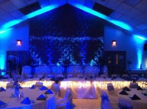 In a realm where elegance and sophistication converge, the banquet hall stands as a beacon of style, awash in the radiant glow of blue and white lighting. Like a superheros cloak, tables are clad in pristine white linens, enhanced by the striking contrast of blue napkins—every detail meticulously planned to create an ambience that inspires awe. Enter Black Tie Productions—the quintessence of event mastery—ready to elevate your occasion to legendary status with seamless execution and lightning-fast price quotes for unparalleled services. Transform your event into an extraordinary affair fit for heroes!. :The Event Heroes at Black Tie Productions