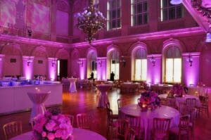 In a majestic ballroom setting where the air crackles with purple luminescence, floral centerpieces herald elegance upon each table. Above, glistening chandeliers dangle like shimmering stars from the cavernous ceiling, while towering arched windows stand guard with grace and grandeur. Transform this exquisite ambiance into a realm of legend with the Event Heroes photo booth rental—a portal to immortalize heroic memories from your unforgettable night!. :The Event Heroes at Black Tie Productions