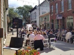 In the heart of the city, where charming brick buildings rise like steadfast guardians, an elegant outdoor street wedding ceremony unfolds. Guests, akin to noble allies, sit poised on white chairs under a canopy of twinkling string lights that dance in alliance with professional event illuminations. Justice and sophistication are served by a powerful duo—a speaker and a laptop from Black Tie Productions—standing vigilant on the left, orchestrating an aura of grace and grandeur as loves mission commences beneath this starry canopy. :The Event Heroes at Black Tie Productions