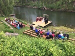 In a realm where love soared as high as the clearest blue sky, an outdoor wedding unfurled by the tranquil river. Mighty wooden benches lined up like valiant guardians, hosting friends and family ready to witness destiny’s embrace. At the heart of this wondrous gathering stood the bride and groom, heroes united under a majestic arch carved from enchanted timber. Their loyal allies—bridesmaids in radiant armor and groomsmen wielding grace—flanked them with unwavering support. Verdant greenery enveloped the scene in a vibrant cloak of life. From above descended Event Heroes, champions known across lands for their legendary photo booth rental services. With dazzling precision and flair, they enhanced this monumental celebration into an event of epic proportions!. :The Event Heroes at Black Tie Productions