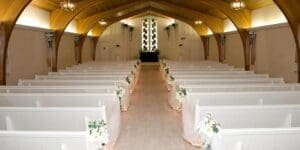 In the heart of this sacred sanctuary, towering wooden beams stretch skyward like mighty pillars of justice. Beneath them, white pews stand at attention, each adorned with blossoms that nod to natures grandeur. At the forefront, a breathtaking stained glass window casts multicolored rays across the room, painting it with an ethereal glow—a testament to artistry and inspiration. An elegant photo booth rental from Event Heroes stands watch as well—its presence a modern marvel—capturing moments of joy and unity within this hallowed haven. :The Event Heroes at Black Tie Productions