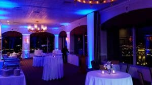 In the heart of the shadowy banquet hall, a powerful glow emanates from blue uplighting, casting an aura of majesty that transforms the space into a realm of elegance. Mighty round tables, draped in pristine white tablecloths and fortified with floral centerpieces, stand as beacons of sophistication. Some are flanked by candles that flicker like eternal flames guiding lost souls to safety. Towering windows serve as portals to a breathtaking panorama of the city’s nighttime skyline—a dazzling metropolis that twinkles under the watchful eyes of vigilant guardians ready to spring into action when duty calls!. :The Event Heroes at Black Tie Productions