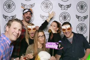 In the heart of a bustling gathering, six vibrant Event Heroes stand united, beaming with joy as they wield their beer-themed props like mighty symbols of camaraderie. Against a backdrop emblazoned with logos and bathed in dynamic, colorful lights, our valiant team strikes an epic pose. This legendary moment captures the essence of celebration and heroism at its finest!. :The Event Heroes at Black Tie Productions