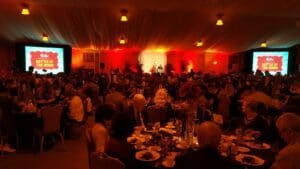 In the grand hall of justice, where heroes and citizens alike gather under a ceiling as vast as the night sky, Black Tie Productions orchestrates an epic gathering. Rows of round tables stretch like tactical conference stations, each occupied by supporters eagerly awaiting the evening’s showdown. Two colossal screens dominate the room with electric energy, proclaiming Battle of The Bands in bright letters that shimmer through a veil of sophisticated lights. This is no mere event; it is a convergence of talent and heart—a harmonious clash for ultimate musical supremacy!. :The Event Heroes at Black Tie Productions
