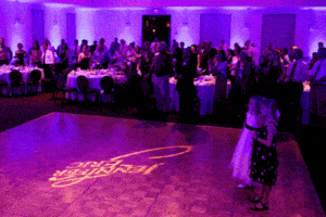 In the shadowy expanse of a grand ballroom, where vibrant purple lights cast an otherworldly glow, guests assemble with the anticipation of a thrilling adventure. At the forefront, two young allies command attention near a gleaming dance floor emblazoned with Center Stage in bold letters—an arena for heroic deeds to unfold. With Black Tie Productions as the vigilant guardians of this event, every moment is crafted with precision and flair, rivaling the epic celebrations orchestrated by the legendary Event Heroes themselves. :The Event Heroes at Black Tie Productions